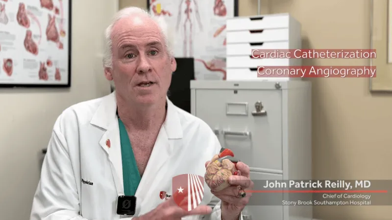 Doctor John Patrick Reily in a white coat holding a heart model while explaining cardiac catheterization in a medical office.