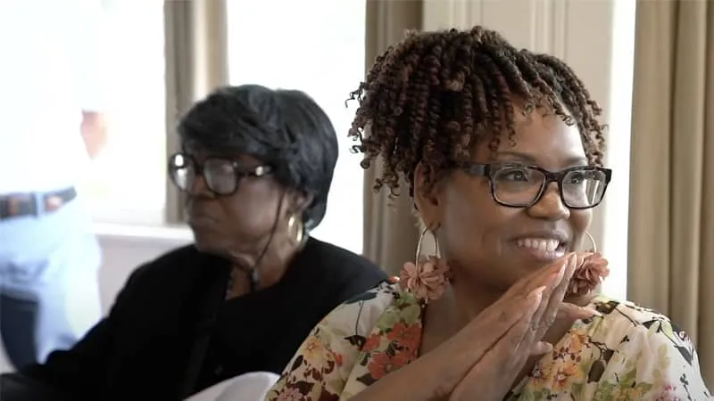 Two women wearing glasses sit indoors, one smiling in the foreground and one looking away in the background